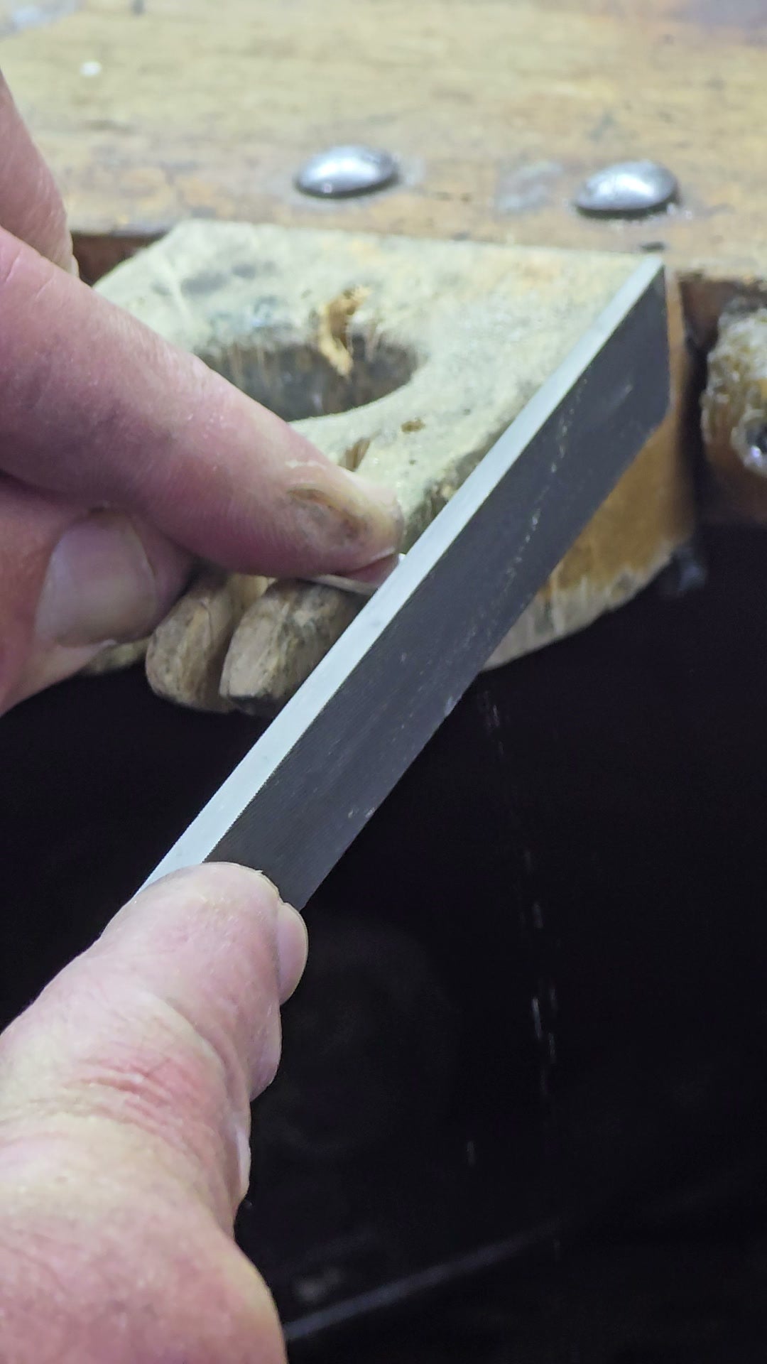 Hands filing a piece of metal against the wooden bench pin, filings falling
